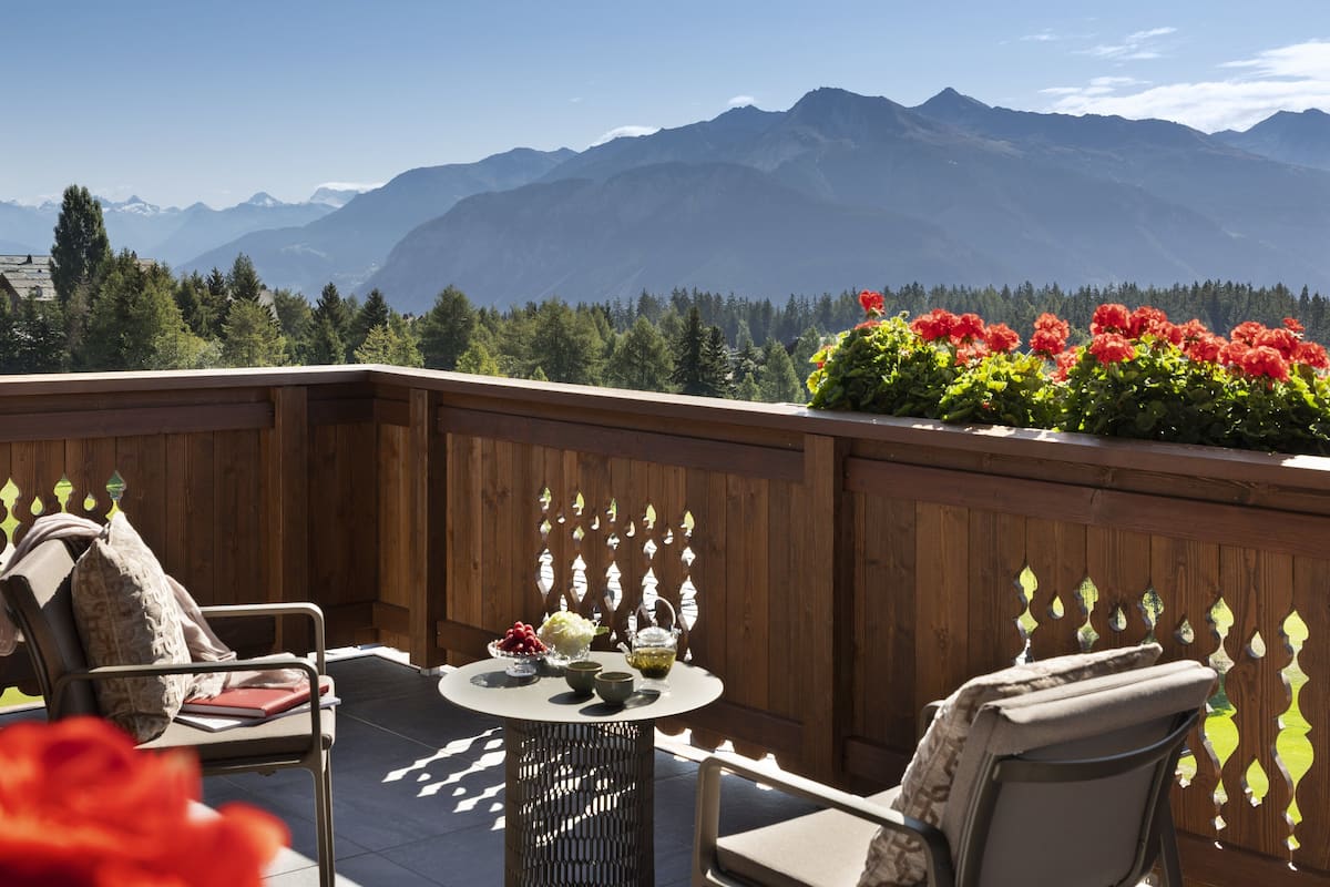 Eine Holzdiele mit einem runden Tisch und Stühlen, ein kleiner Garten mit roten Blumen und Blick auf die Berge.