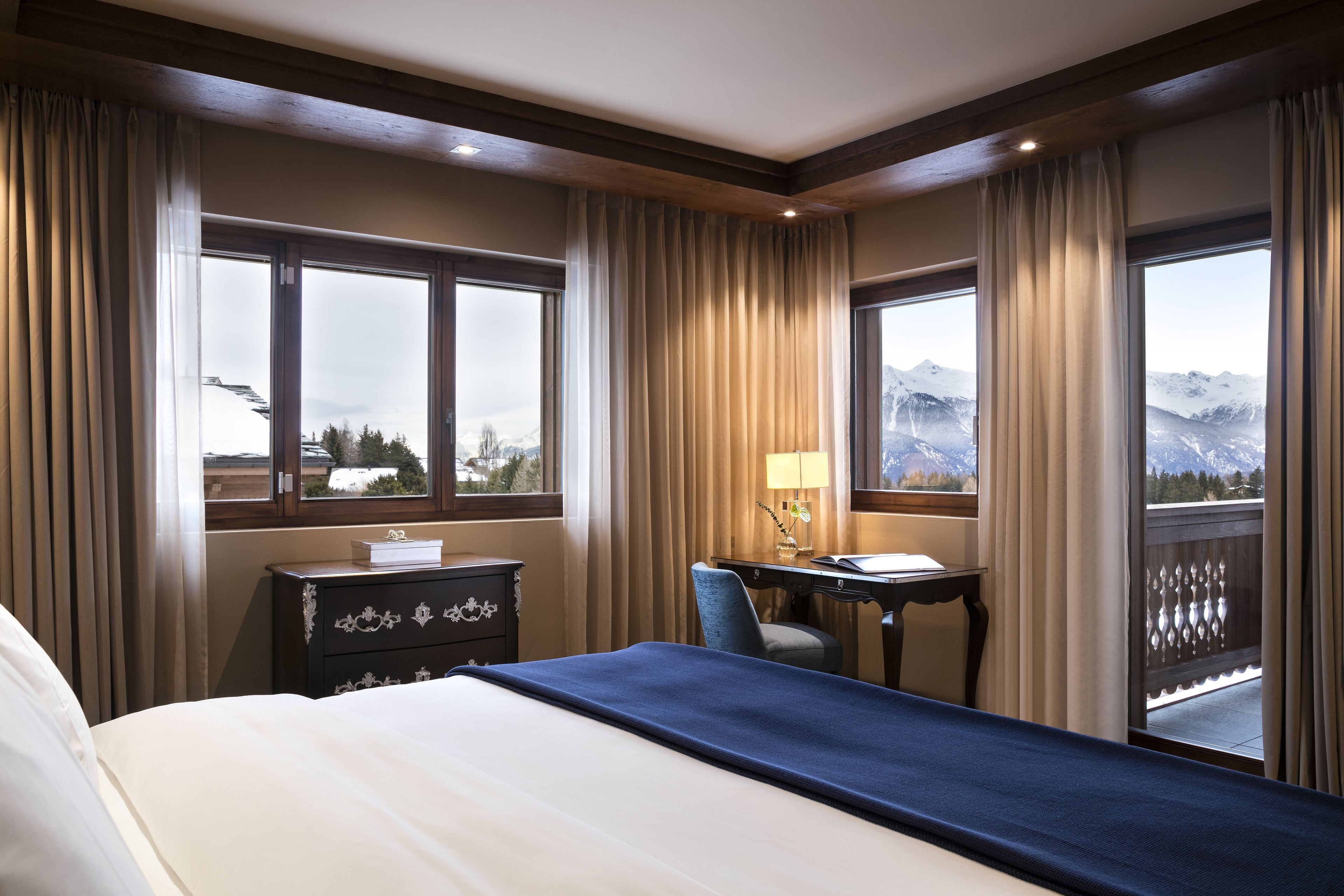 A hotel room with a large bed, a desk with a chair, and a view of mountains through the windows.