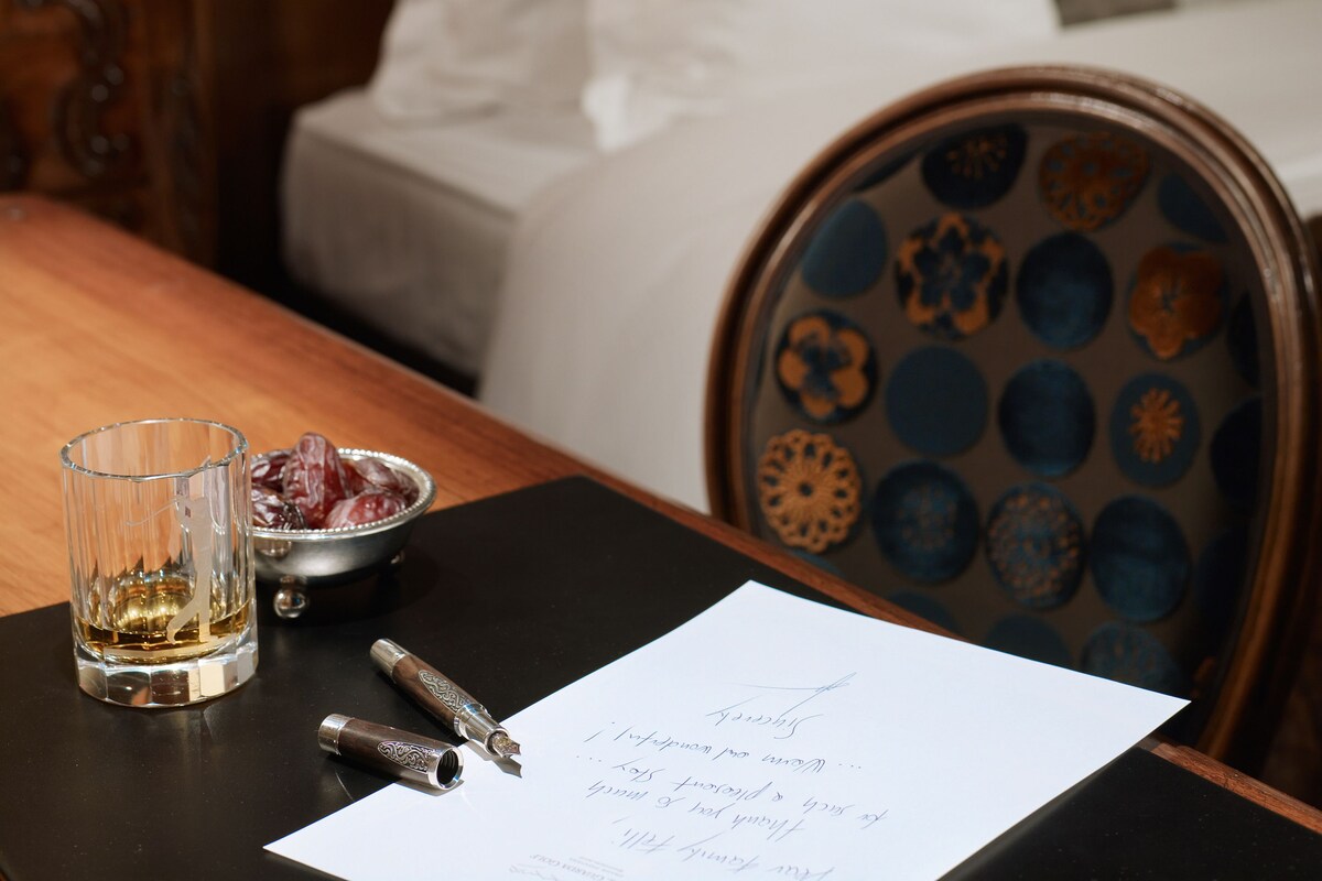 A wooden bed with white bedding, a glass table with a pen and a glass of whiskey, and a paper with handwritten text.