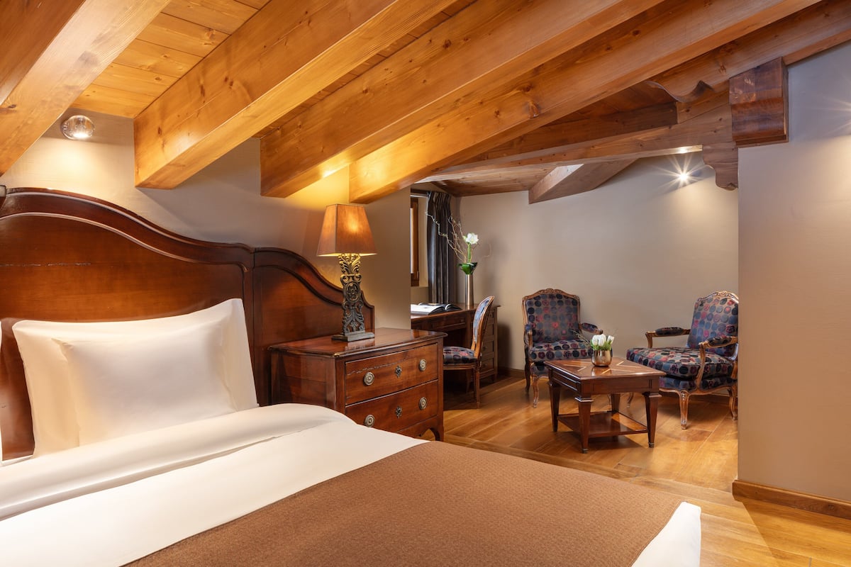 Ein Schlafzimmer mit einem Holzkopfende, einem Holzschrank, einem Schreibtisch, zwei gemusterten Stühlen und einem Spiegel.