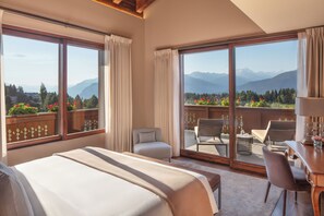 Quarto de hotel com uma cama grande, área de estar com mesa e cadeiras, e varanda com vista para montanhas e flores.