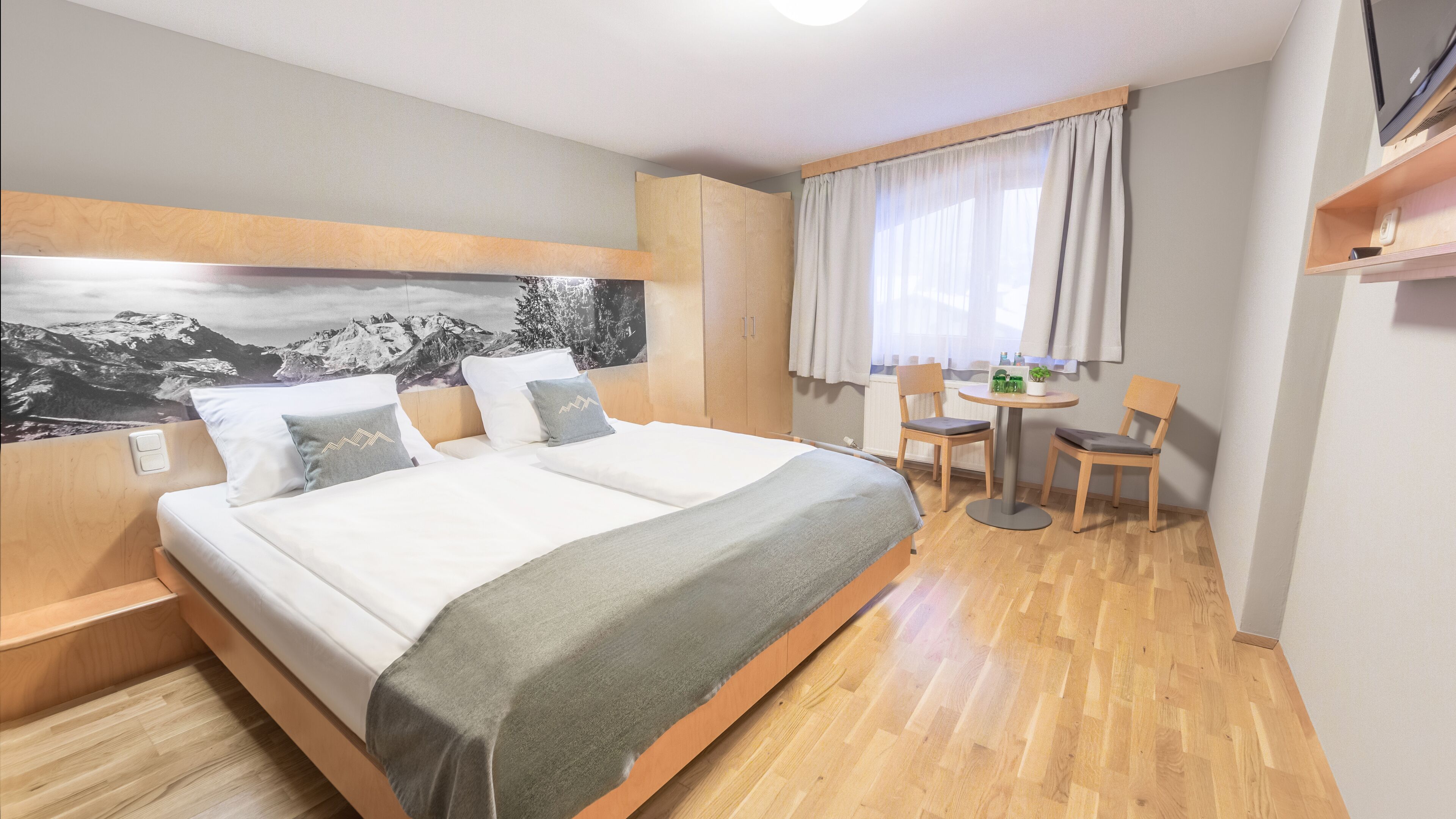 A hotel room with a large bed, a desk, and chairs, a window with curtains, and a wall-mounted TV.