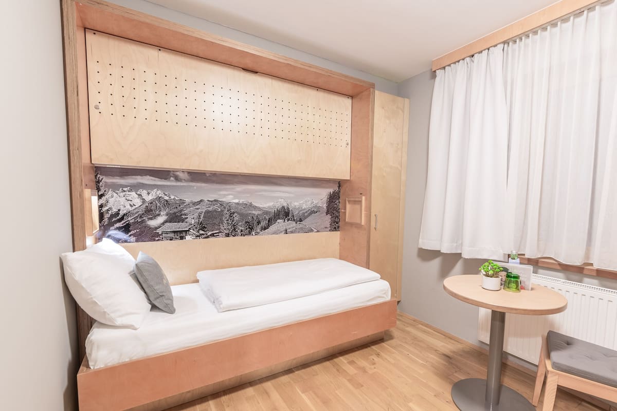 A room with a bed, a small table, a chair, a window with curtains, and a wall-mounted picture.