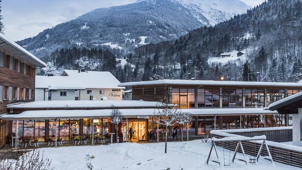 Un hotel con recepción, asientos exteriores y montañas al fondo.