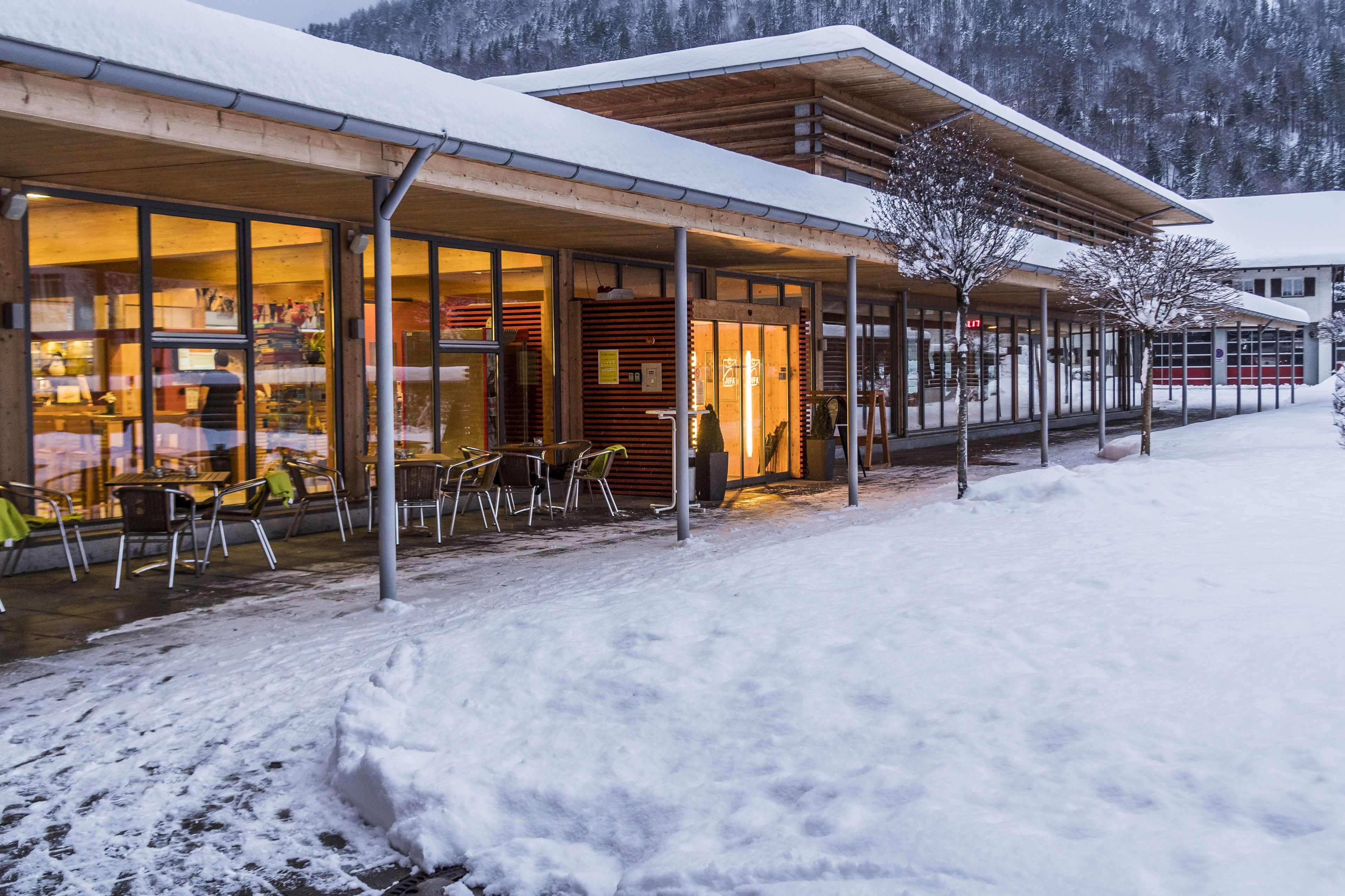 A modern building with large glass windows, outdoor seating, and a snowy landscape.