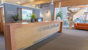 Una recepción con mostrador de madera y pantalla digital, rodeada de plantas en macetas y una vista de paisaje montañoso a través de un amplio ventanal.