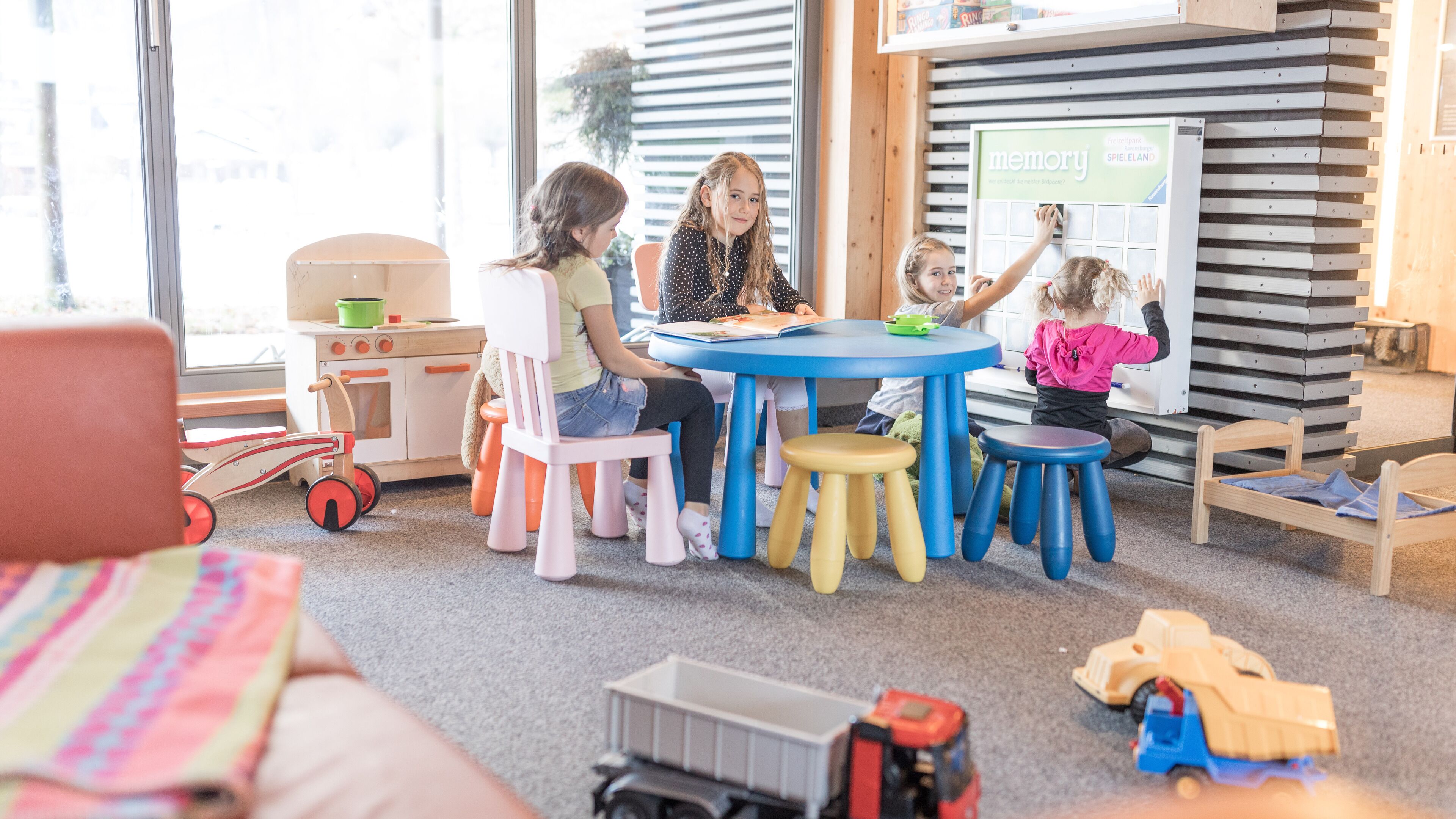 A group of children and an adult at a table with a blue tablecloth, surrounded by colorful stools and a play kitchen setup.