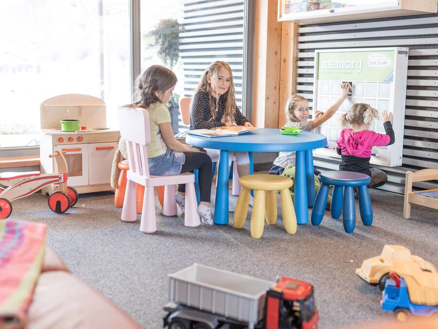 Un grupo de niños y un adulto sentados a una mesa con mantel azul, rodeados de taburetes coloridos y una cocina de juguete.