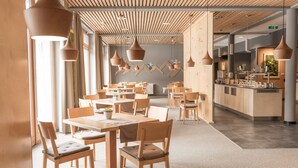 Un interior de restaurante moderno con mesas y sillas de madera, iluminación colgante y una zona de cocina visible al fondo.