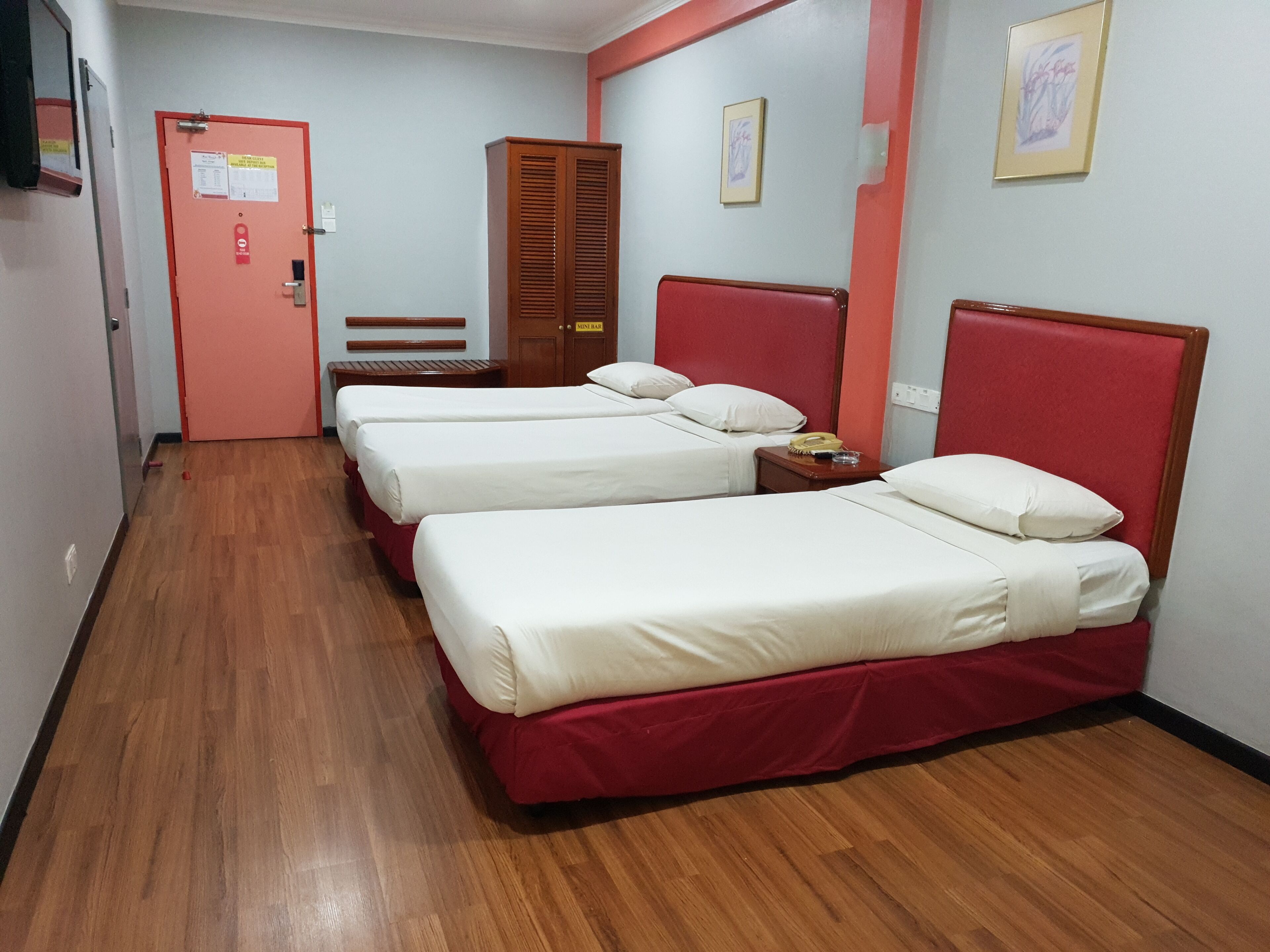 Habitación de hotel con tres camas individuales, suelo de madera, cabecera roja y armario de madera.