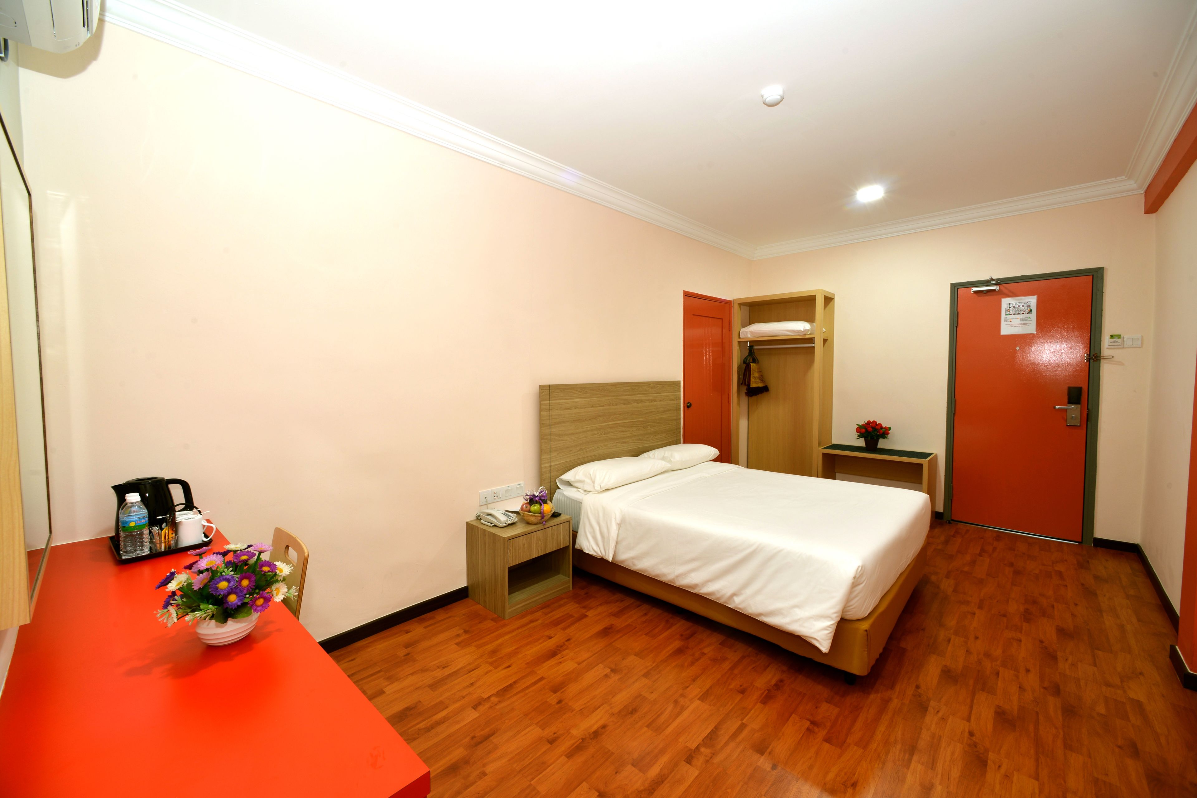 A hotel room with a wooden floor, a bed with white bedding, a desk with a vase of flowers, a nightstand with a phone, and a red door.