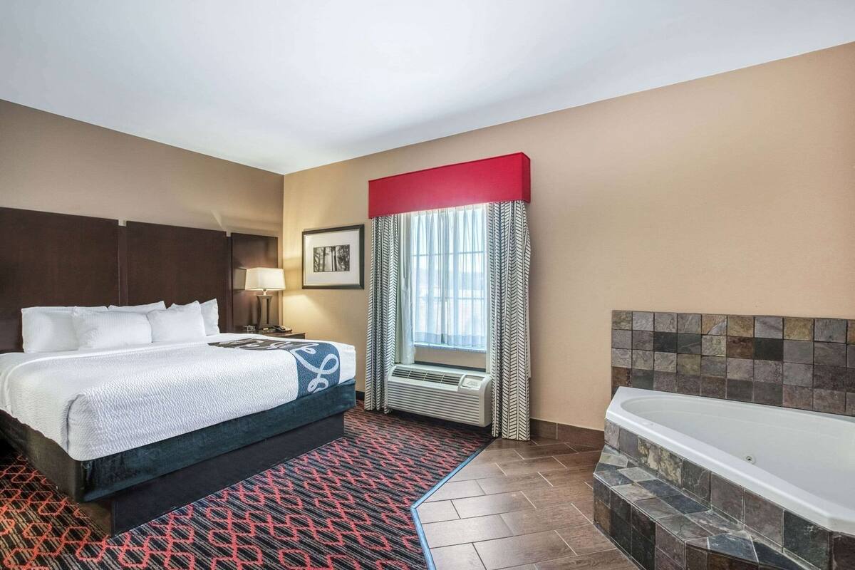 Habitación de hotel con una cama grande, baño independiente con bañera, ventana con cortinas e imagen en la pared.
