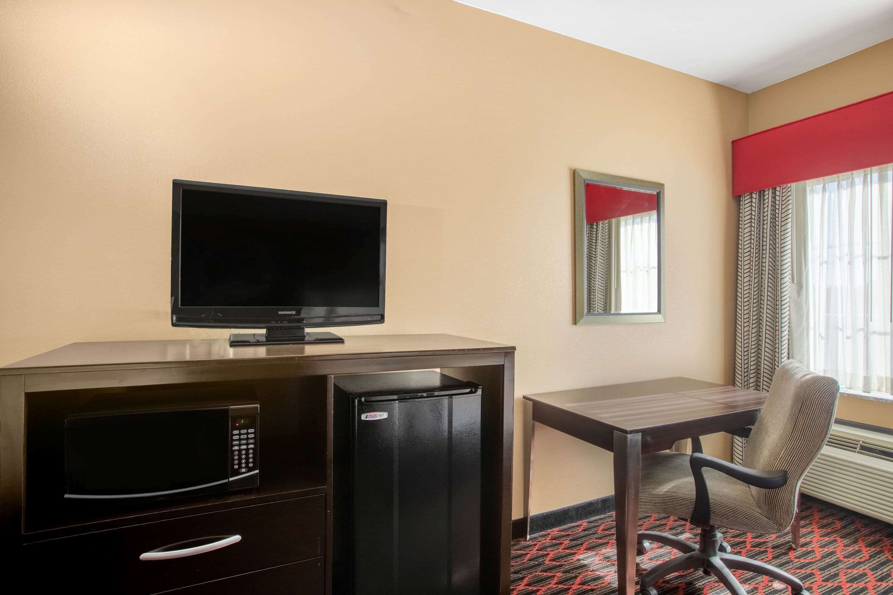 Ein Hotelzimmer mit Schreibtisch, Fernseher, Kühlschrank, Mikrowelle, Stuhl und einem Fenster mit Vorhängen.