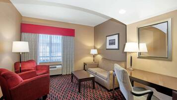 A hotel room with a red armchair, a sofa, a small table, and a desk. There are two lamps, a framed picture on the wall, and a window with curtains.
