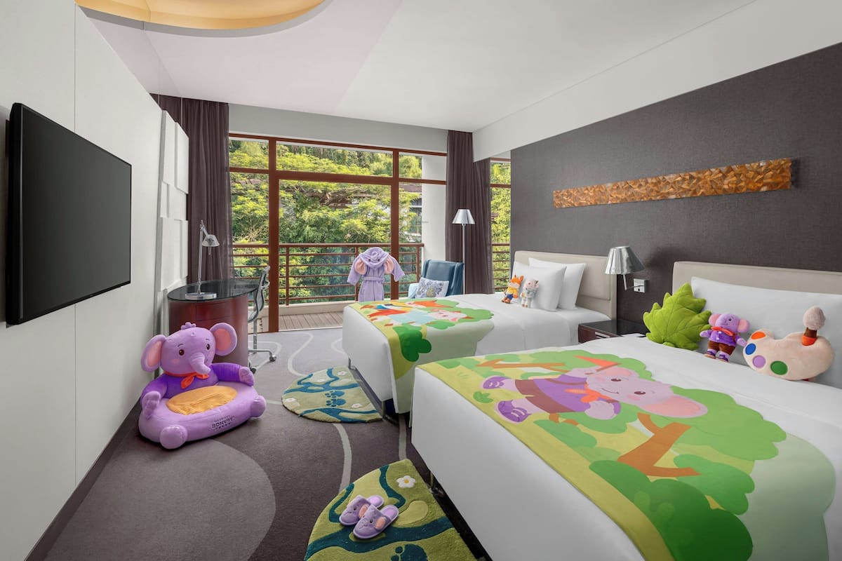 A hotel room with two beds, a TV, a balcony with a view of greenery, and a children's play area with a purple elephant toy.
