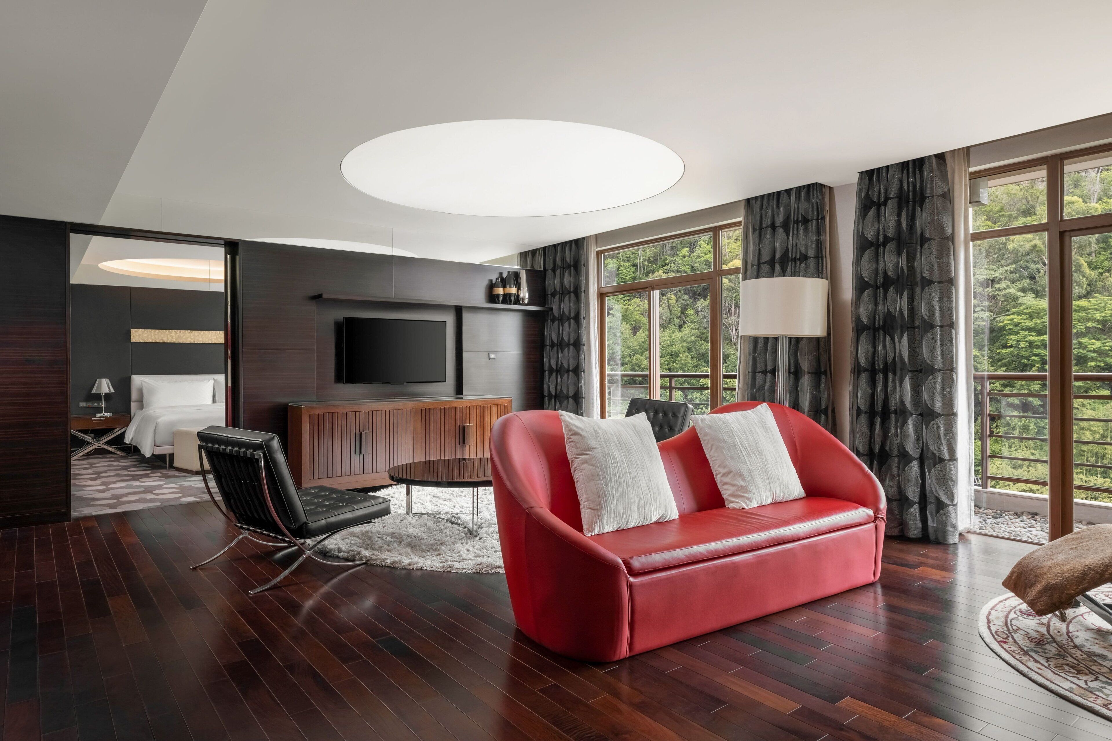 A modern living room with a red leather sofa, a flat-screen TV, and a large window with curtains.