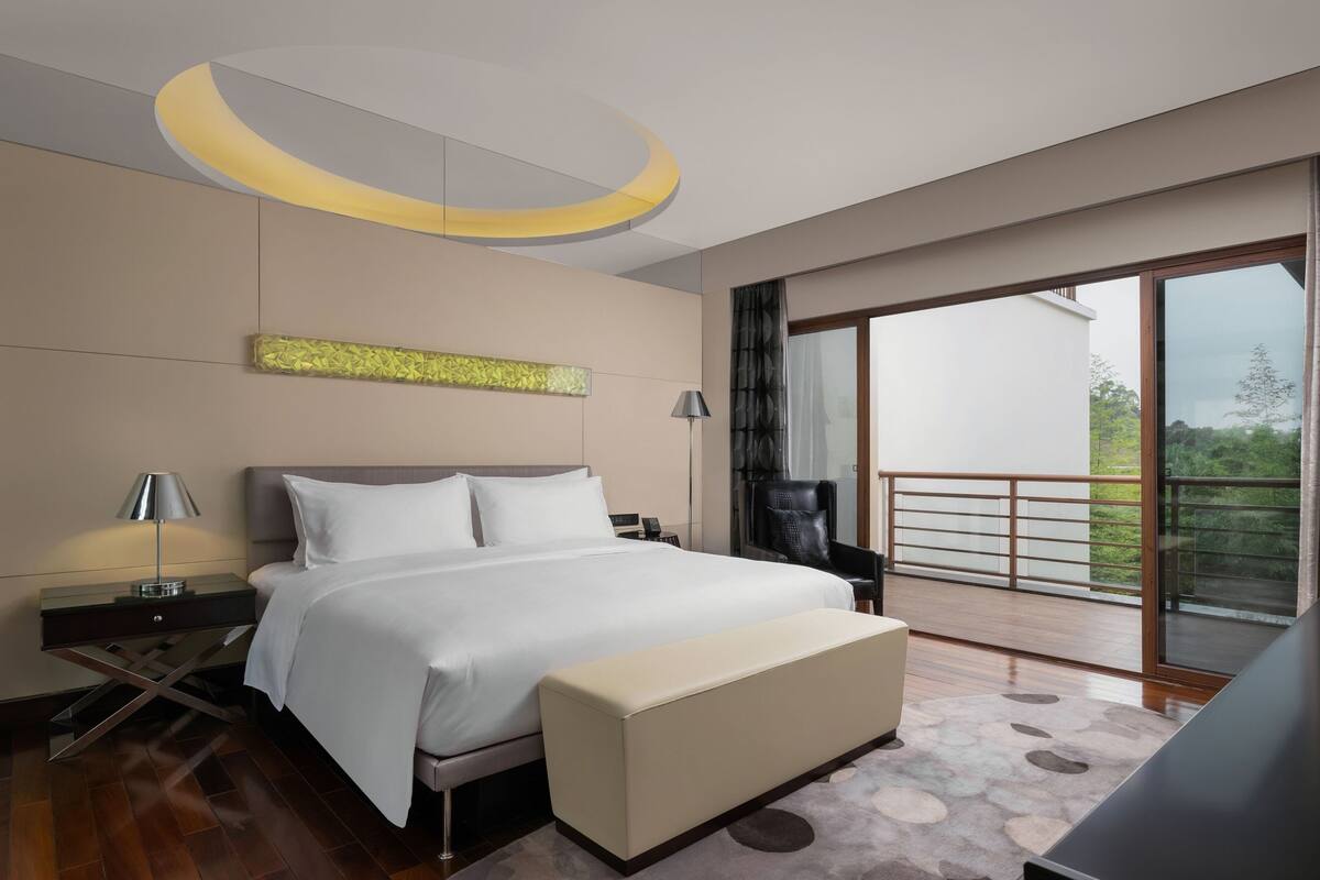 A modern hotel room with a large bed, a bench, a desk, and a balcony with a view.