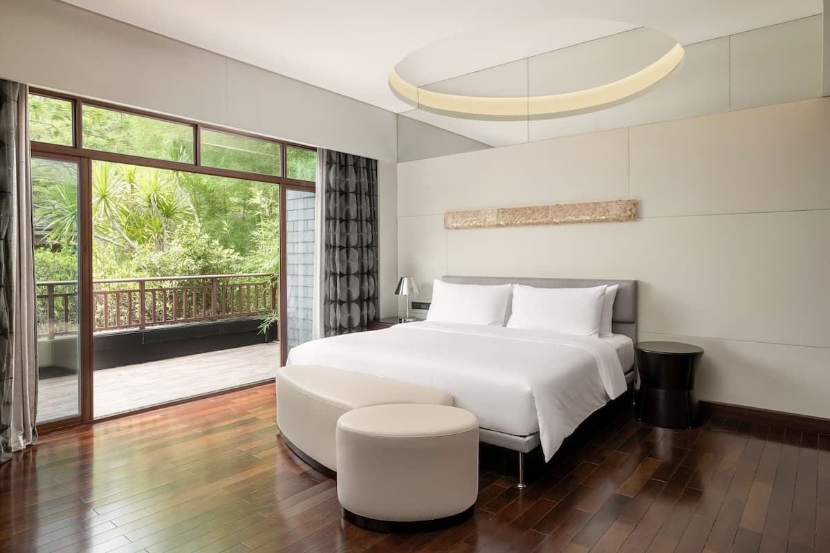 A modern bedroom with a large bed, a sofa, a bedside table, and a view of greenery through large windows.