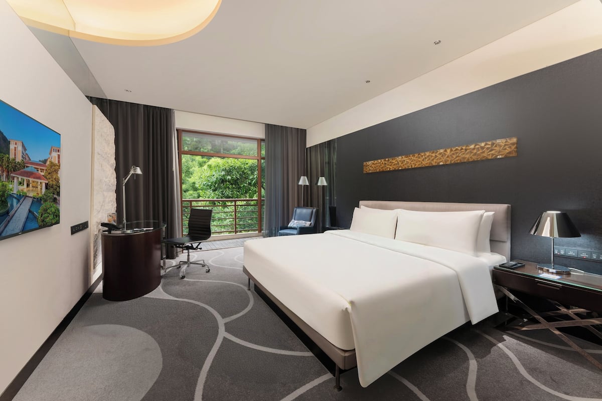 A modern hotel room with a large bed, a desk, and a view of greenery outside.