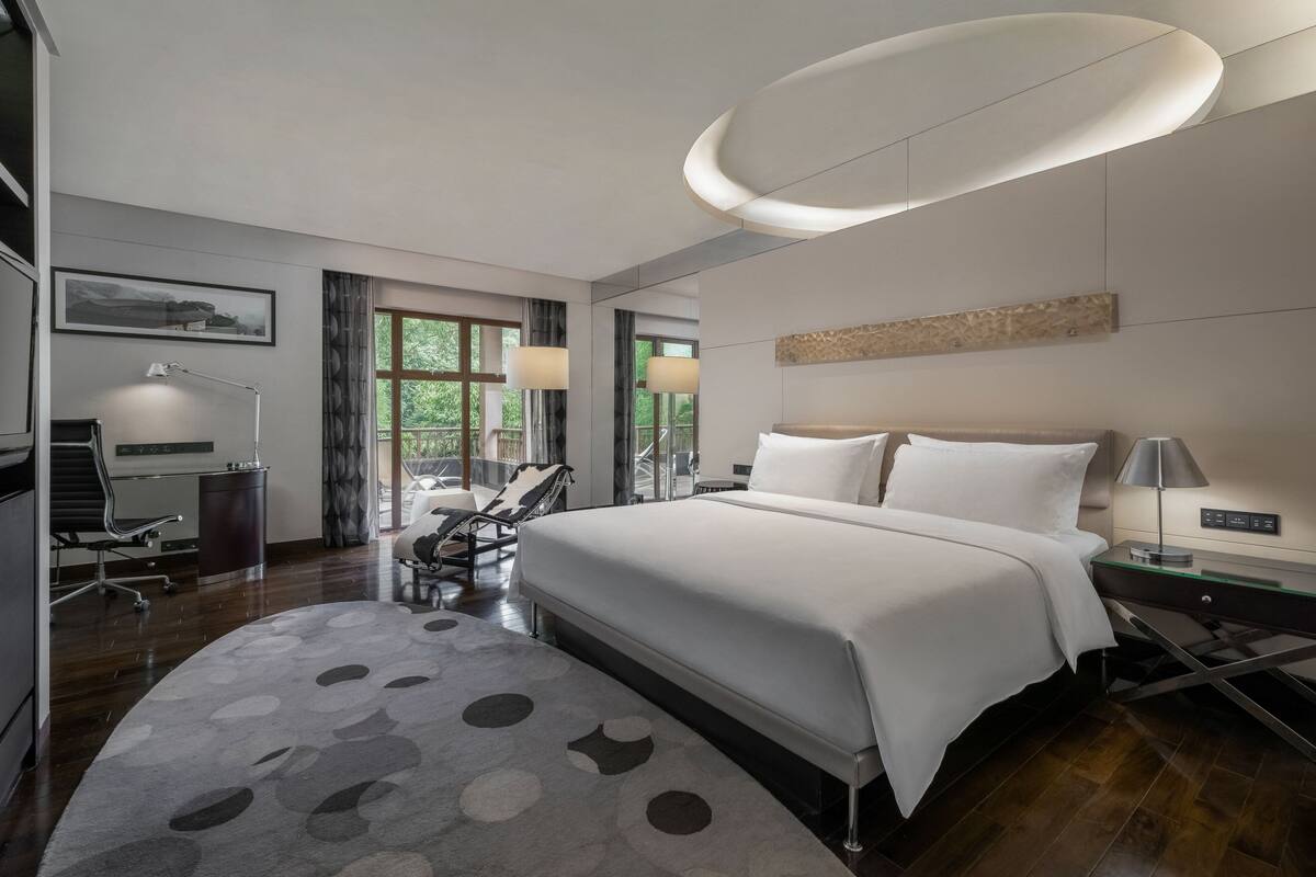 A modern hotel room with a large bed, a desk, a chair, and a view of greenery through the windows.