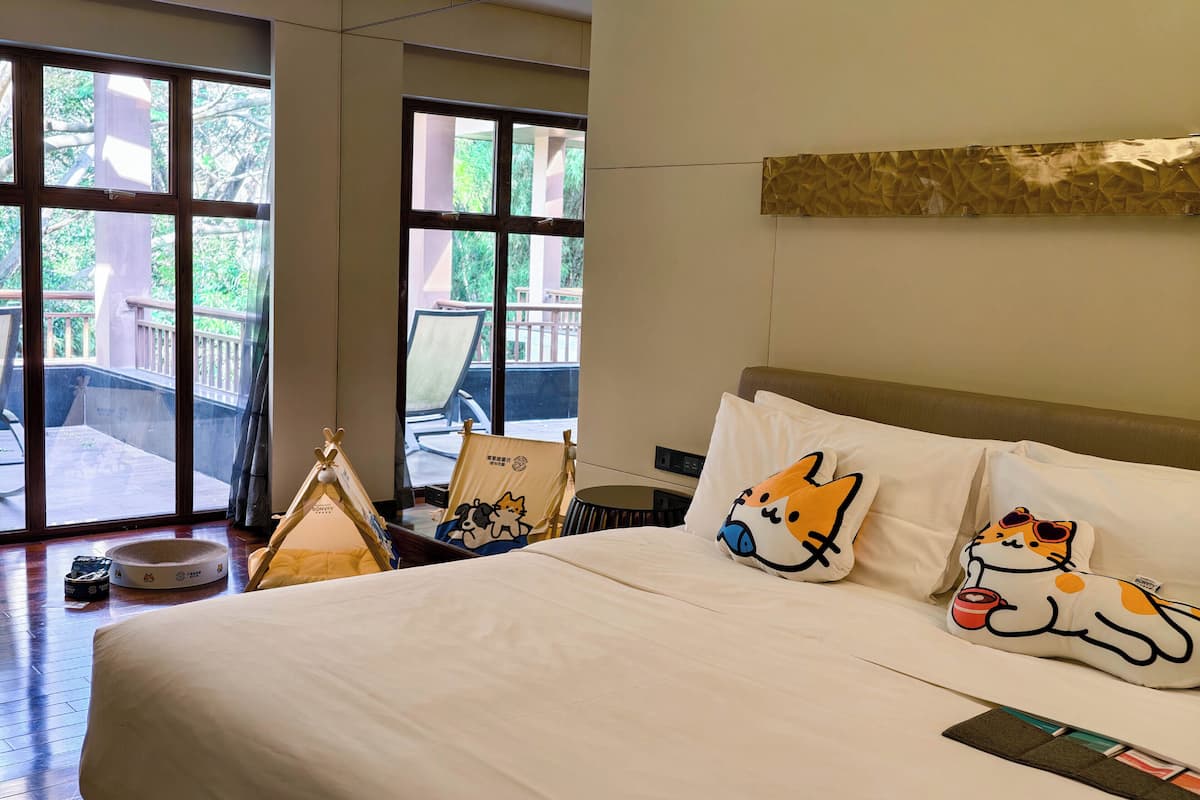 A hotel room with a large bed, two pillows with cat designs, a bedside table, and a view of a balcony with chairs and a table.