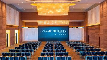 A conference room with rows of tables and chairs, a large screen displaying 'Le Meridien Xiamen', and a chandelier.