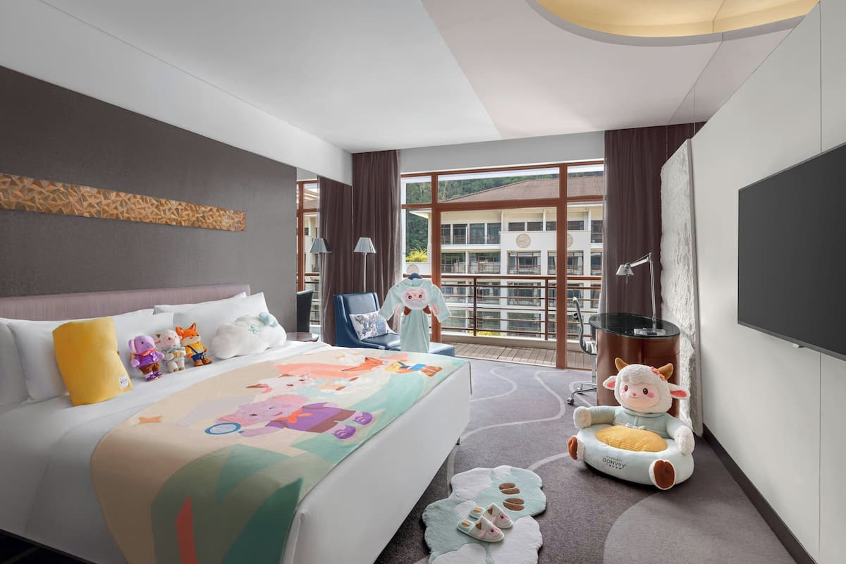 A hotel room with a large bed, a plush toy, a small table, and a view of a building through the window.