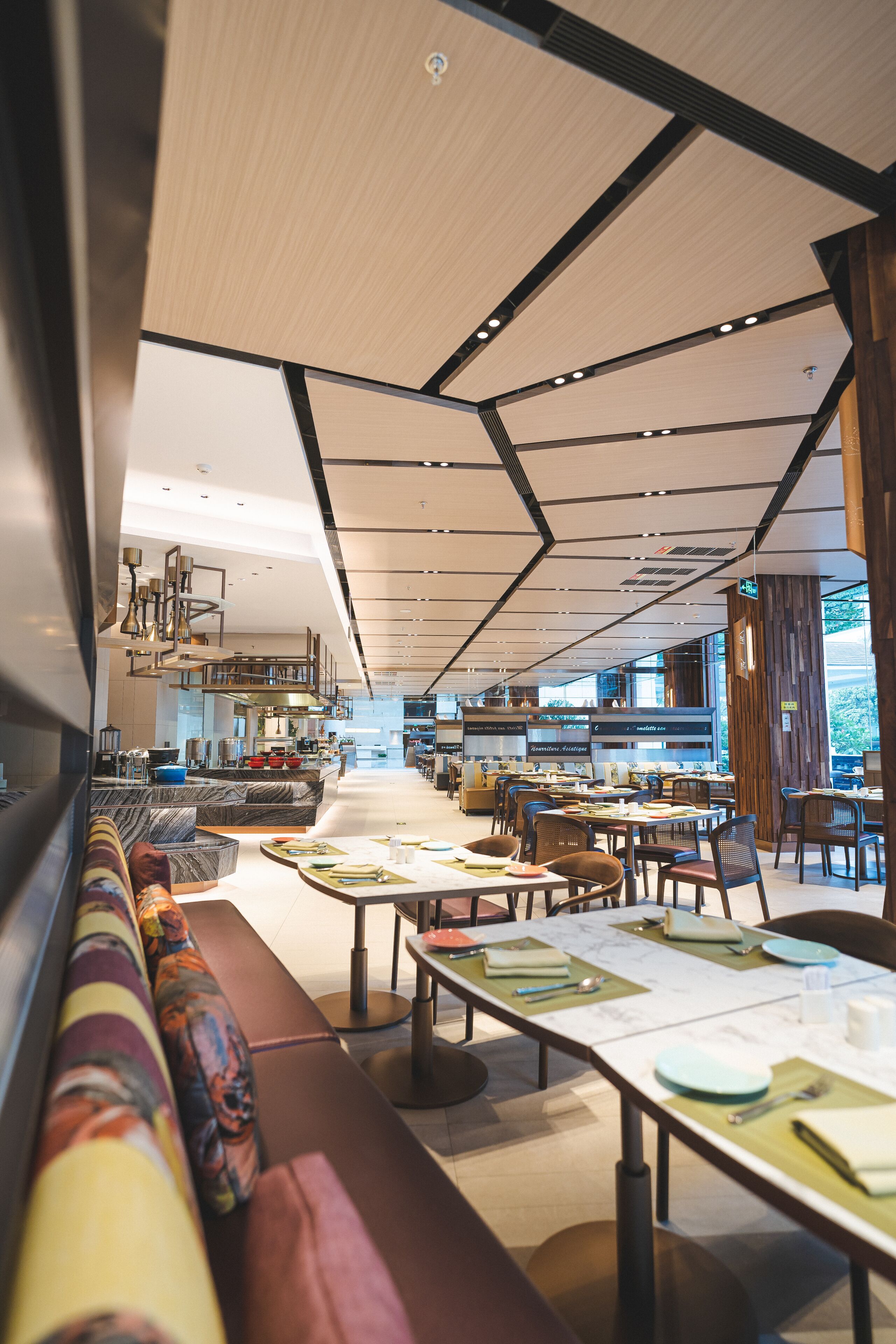 A modern restaurant with a long dining area, a kitchen in the background, and a view of the outdoors.