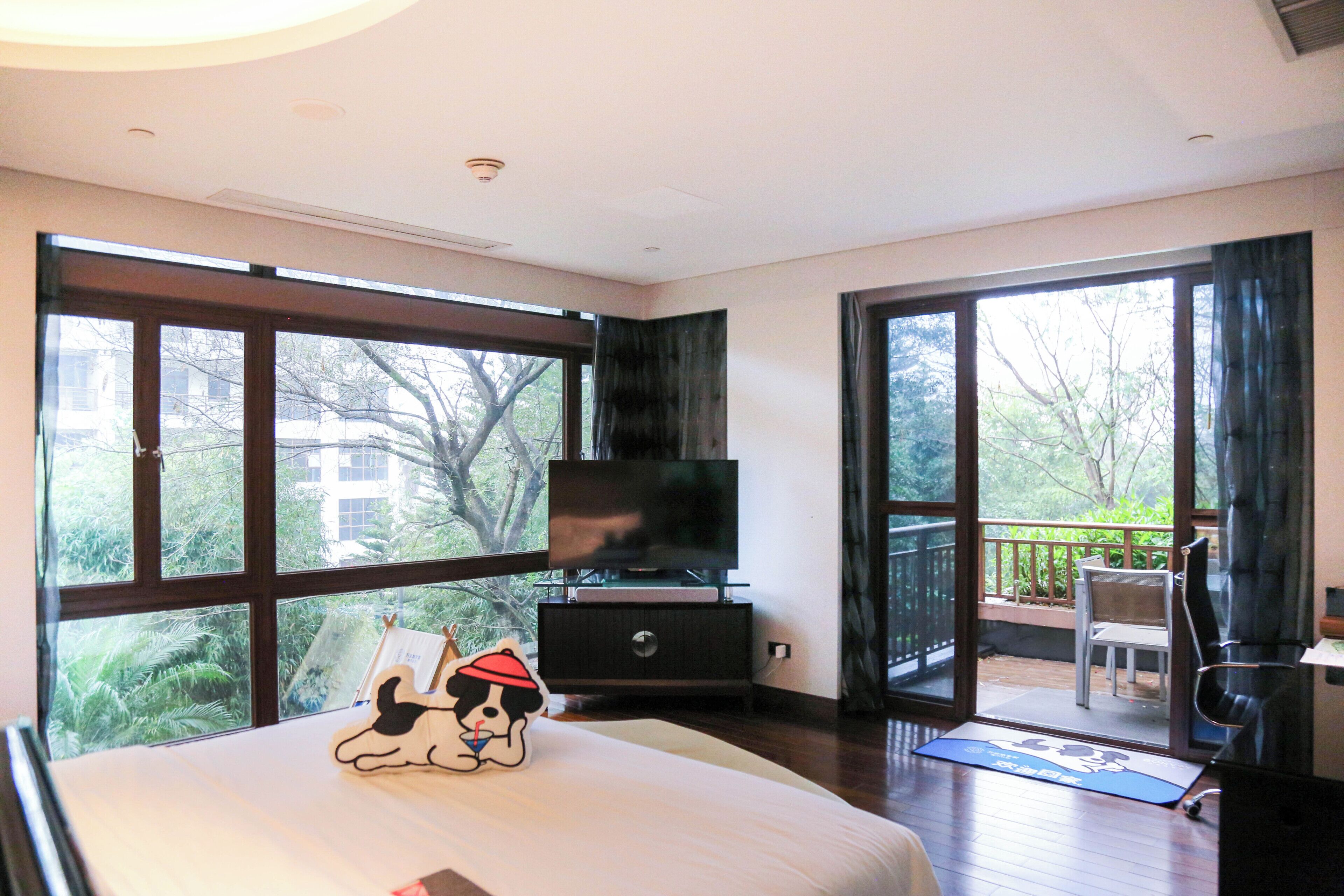 A hotel room with a large bed, a TV on a stand, a balcony with a chair, and a view of trees and buildings.