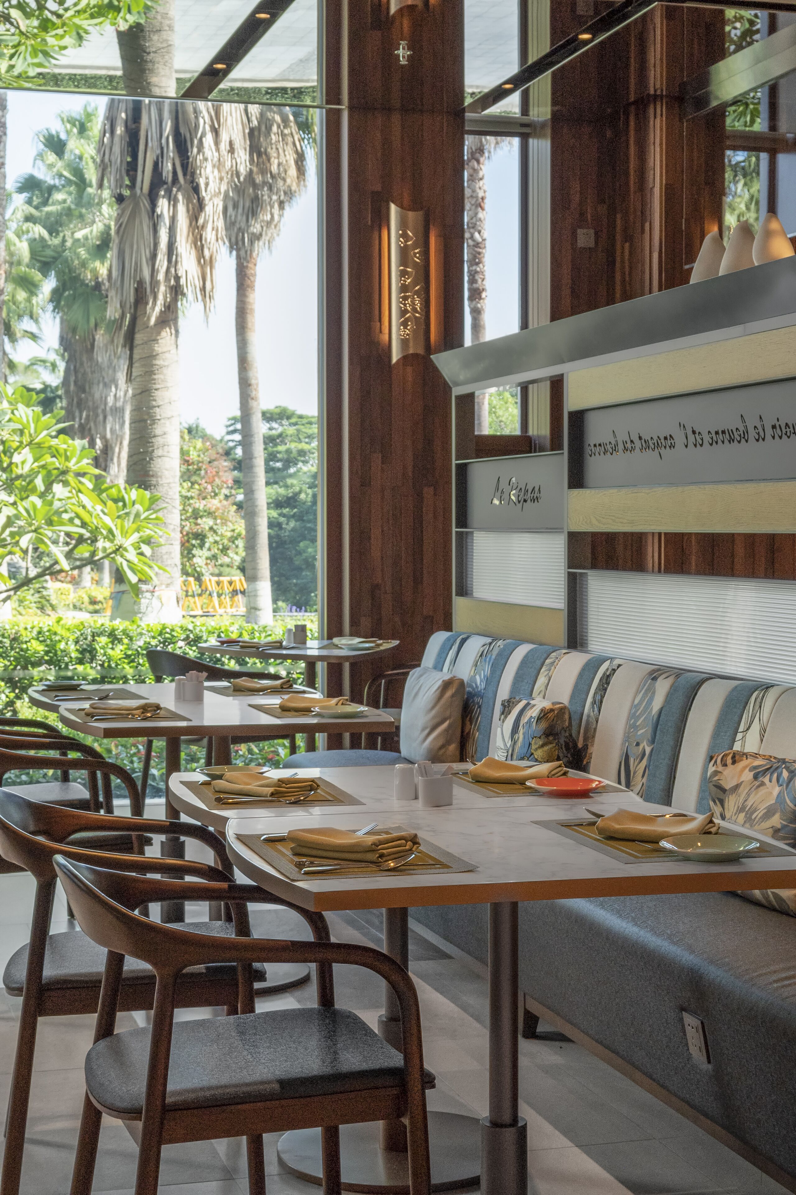 Un restaurant moderne avec des meubles en bois, un canapé à motifs floraux et une vue sur les palmiers, visible à travers de grandes fenêtres.