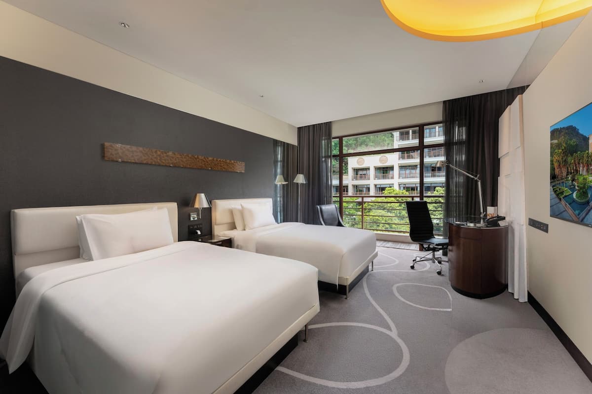 A hotel room with two beds, a desk, a chair, a lamp, and a view of a building through large windows.