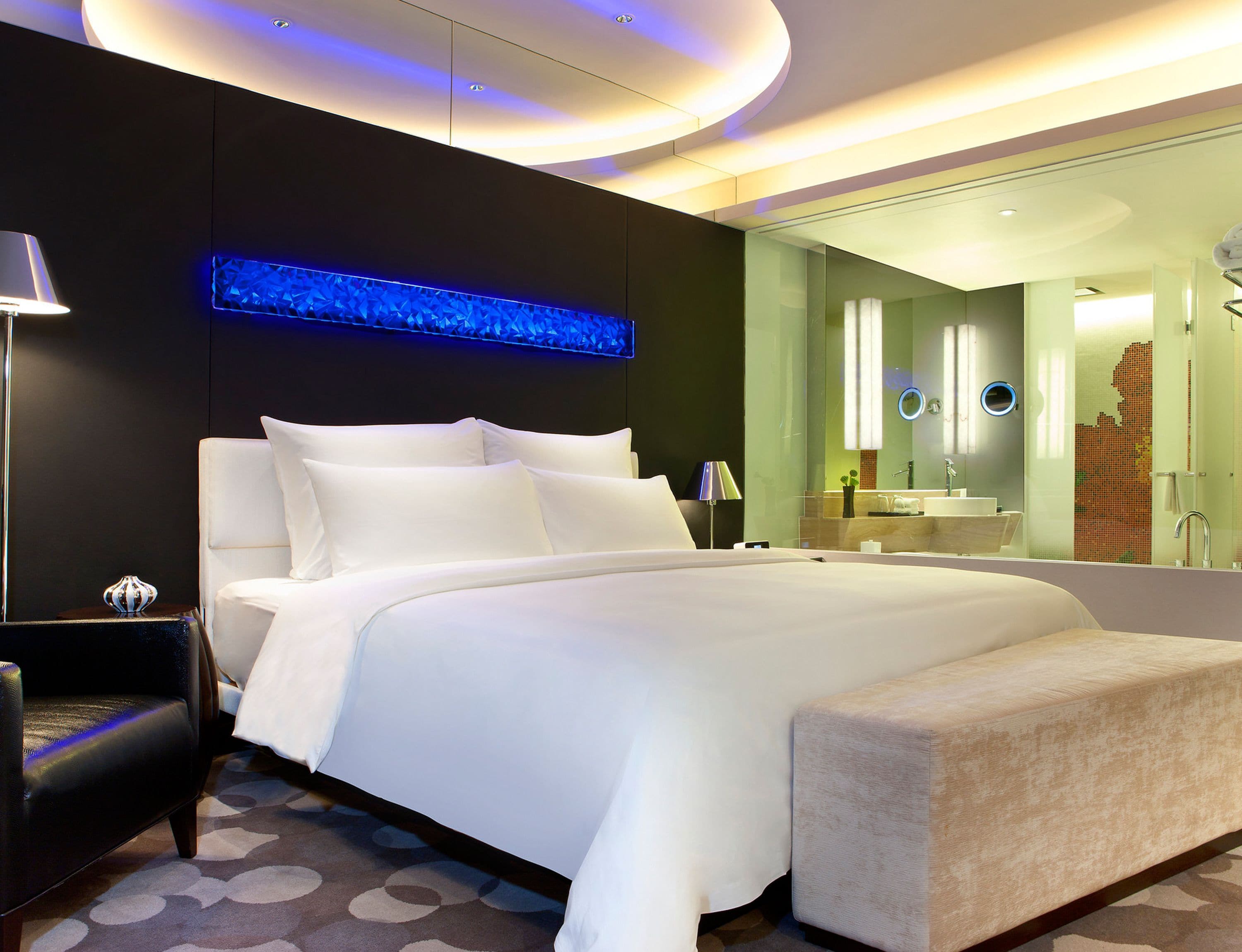 A modern hotel room with a large bed, a bedside table, a lamp, a mirror, and a bathroom with a sink and mirror.
