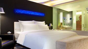 A modern hotel room with a large bed, a bedside table, a lamp, a mirror, and a bathroom with a sink and mirror.