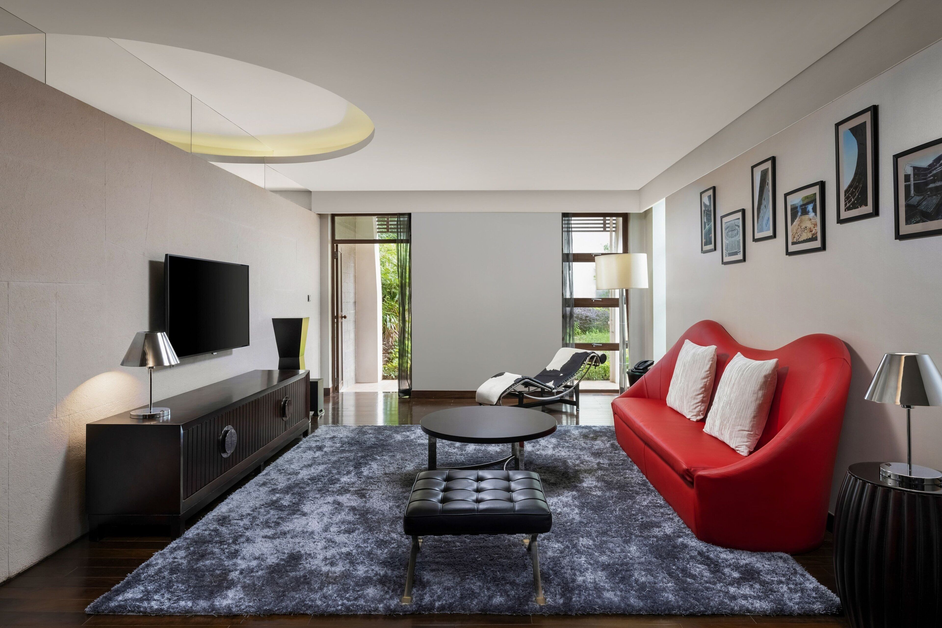 Un salon moderne avec un téléviseur à écran plat, un canapé rouge, un pouf noir et une table basse ronde. Des tableaux encadrés ornent le mur, et une grande fenêtre offre une vue sur la verdure.
