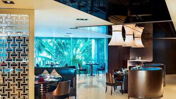 Un hall d’hôtel moderne doté d’un espace salon central, d’un bar et d’une grande fenêtre offrant une vue sur la végétation tropicale.