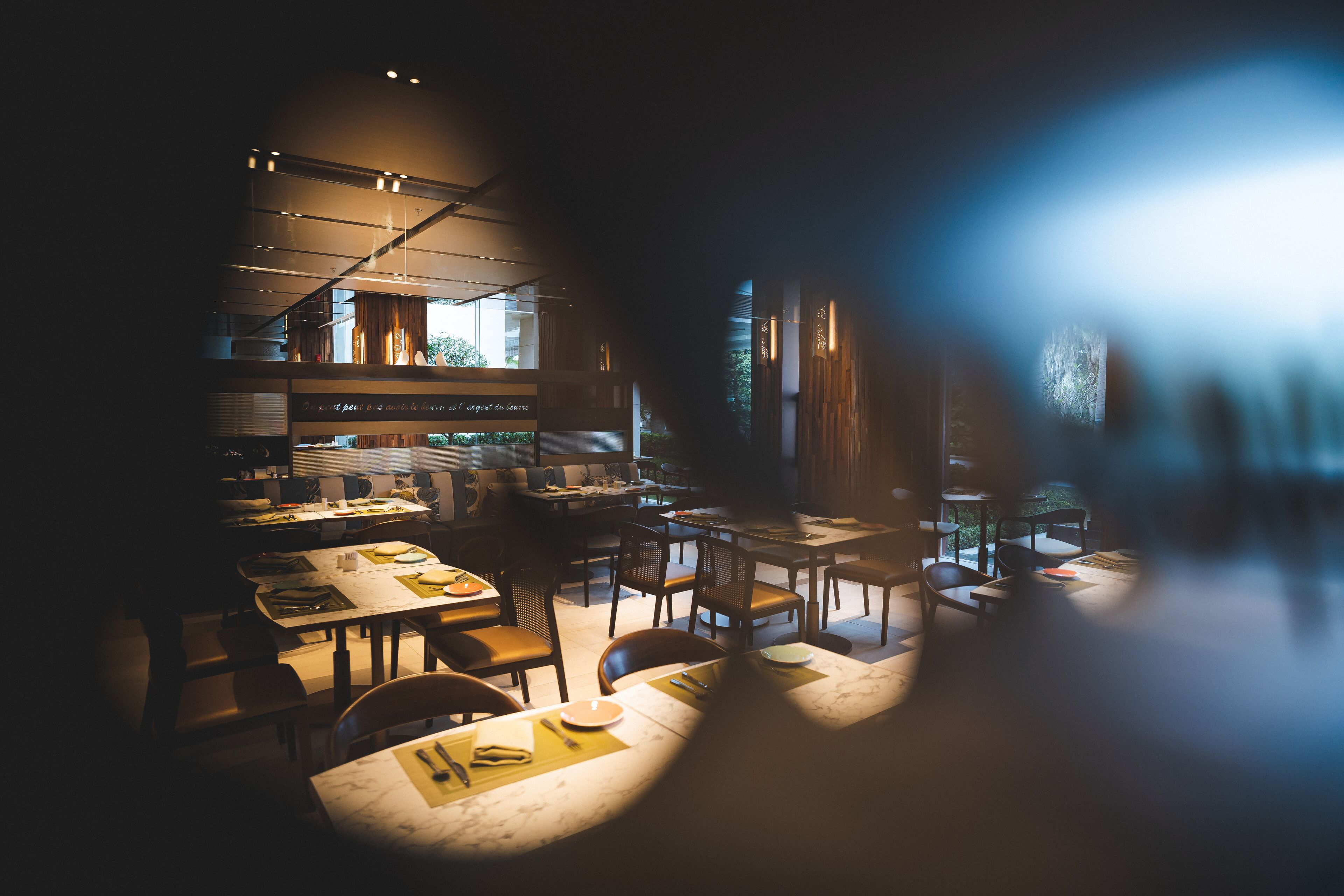 A modern restaurant with multiple dining tables and chairs, a kitchen area, and large windows allowing natural light.