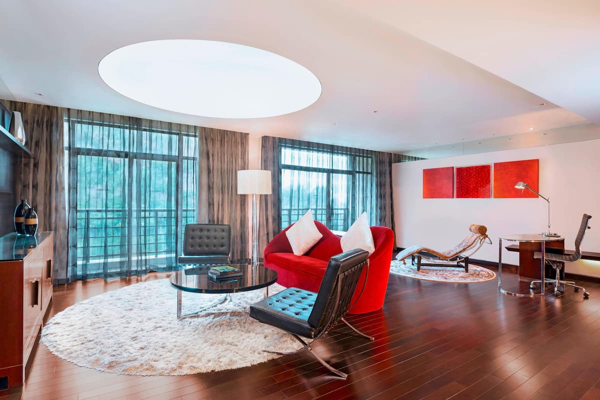 A modern living room with a red sofa, a black leather chair, and a glass coffee table. There is a large window with sheer curtains, a round rug, and a chandelier.
