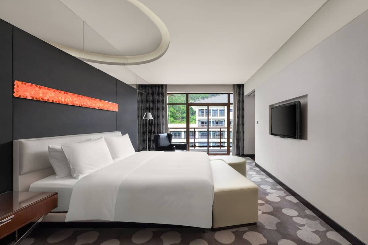 A modern hotel room with a large bed, a desk, a chair, a TV, and a window with a view of buildings.