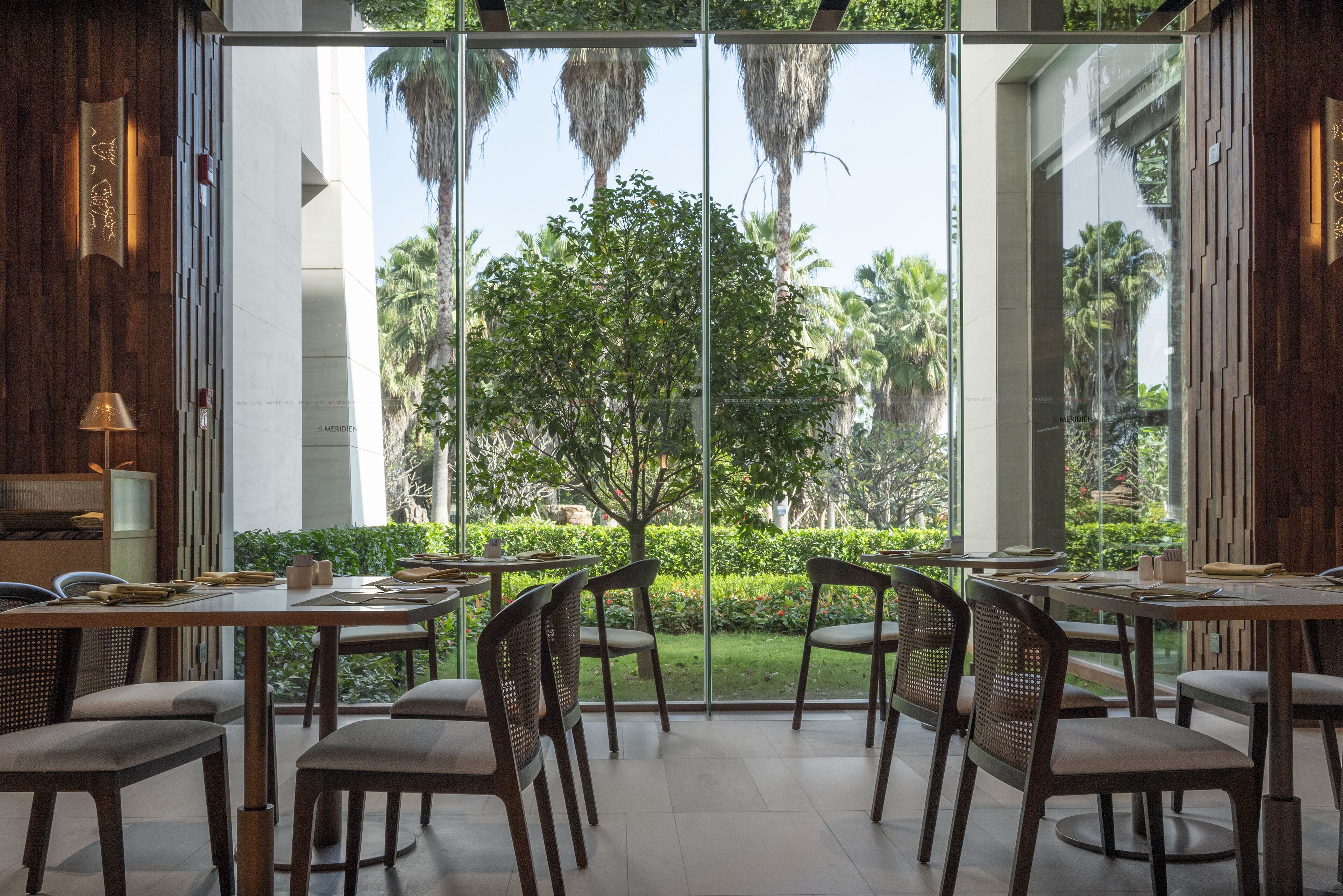 Un comedor moderno con paredes de vidrio, mobiliario de madera y vistas a palmeras.