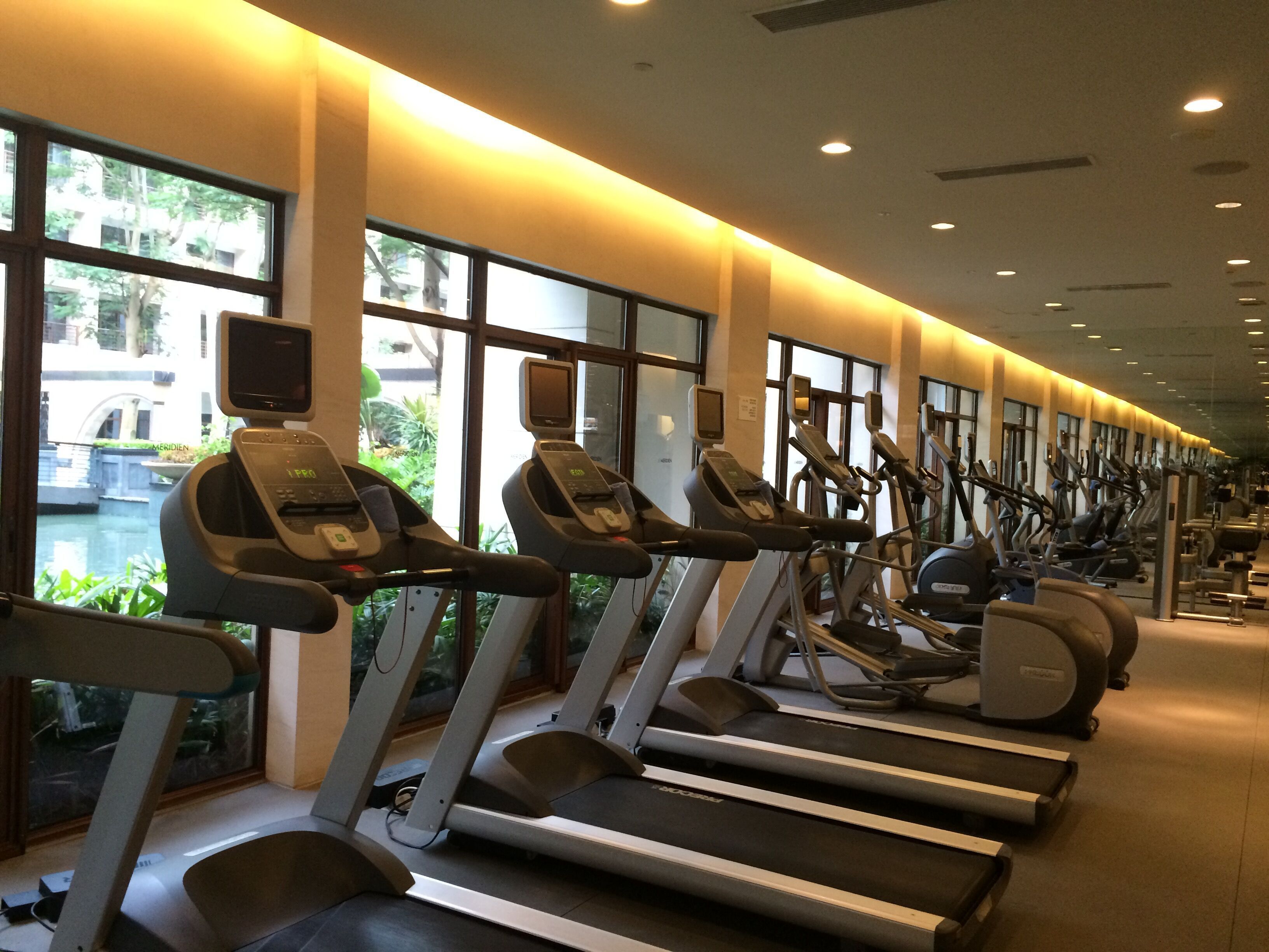 A row of treadmills in a gym with large windows and a view of a building outside.