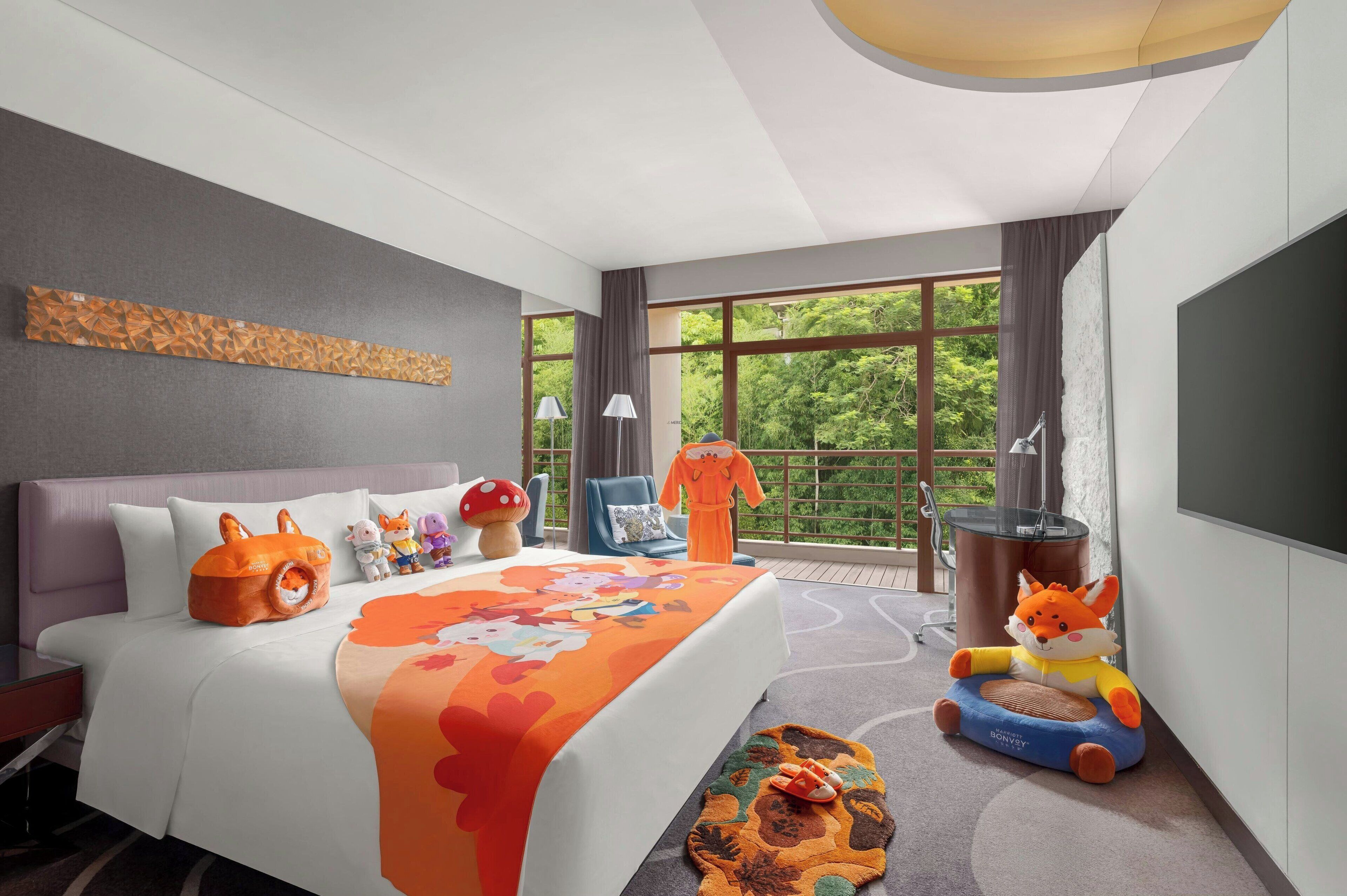A modern hotel room with a large bed, a sofa, a dining table, and a view of greenery through large windows.