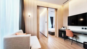 A modern hotel room with a wooden panel wall, a large flat-screen TV, a sofa, a desk with a chair, and a bed in the background.