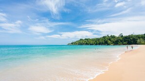 Ein Strand mit klarem türkisblauem Wasser, einem Sandufer und einem bewaldeten Hügel im Hintergrund.