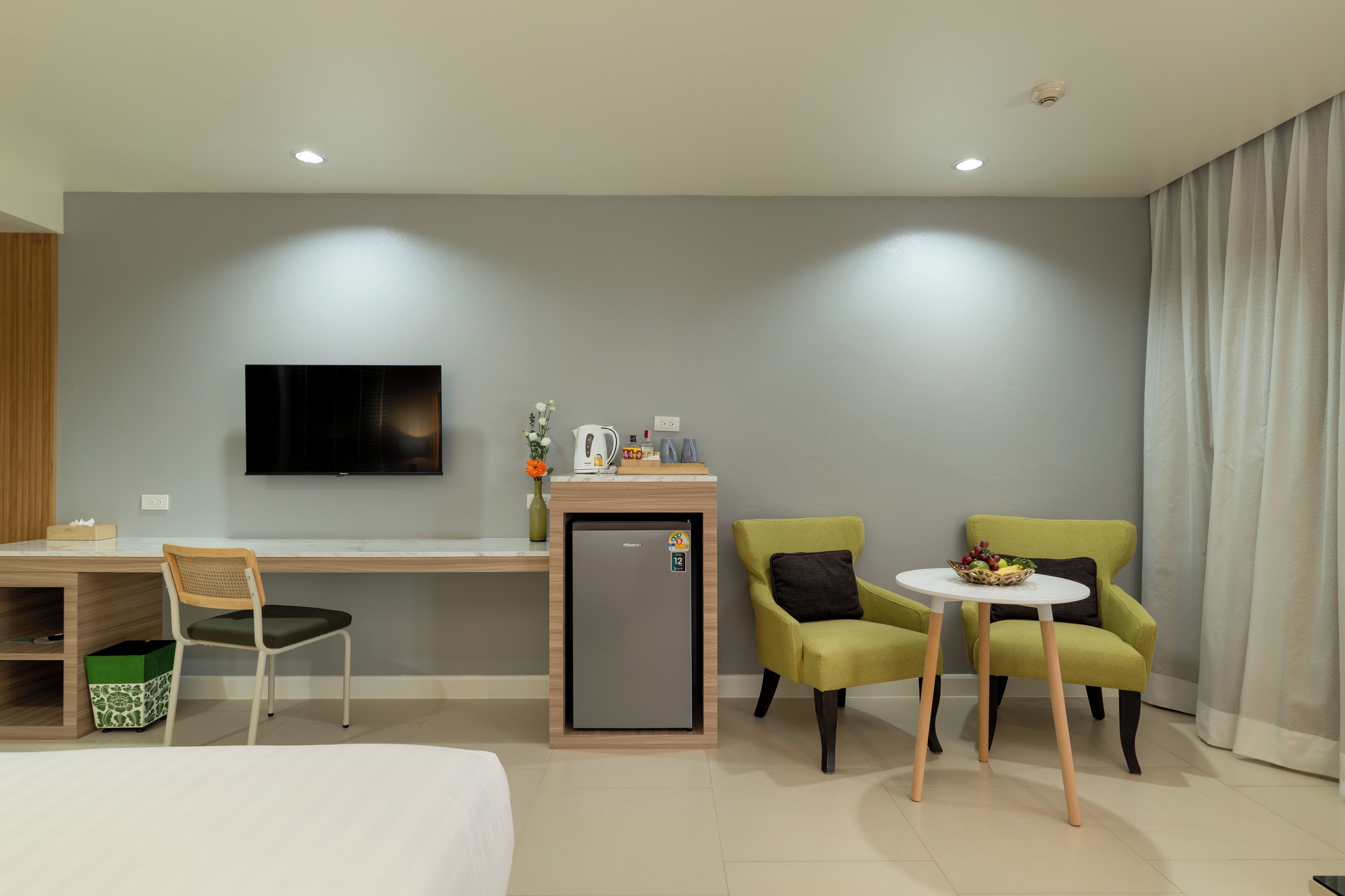 Habitación de hotel con cama, escritorio con televisor, refrigerador pequeño, dos sillones verdes y una mesita con frutero.