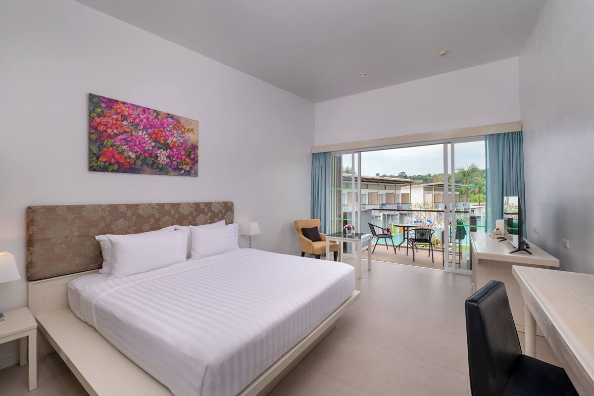 A hotel room with a large bed, a desk, a chair, and a balcony with a view of the city.