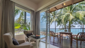 Ein Zimmer mit Blick auf den Strand und Palmen, ausgestattet mit einer Couch, einem Tisch und einem Balkon mit Holzstühlen.