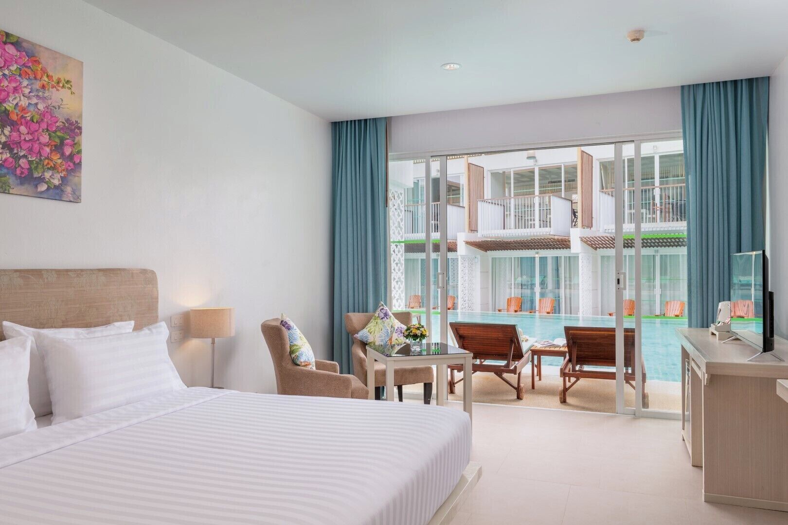 A hotel room with a bed, a desk with a TV, a balcony with a pool, and a view of the building's exterior.