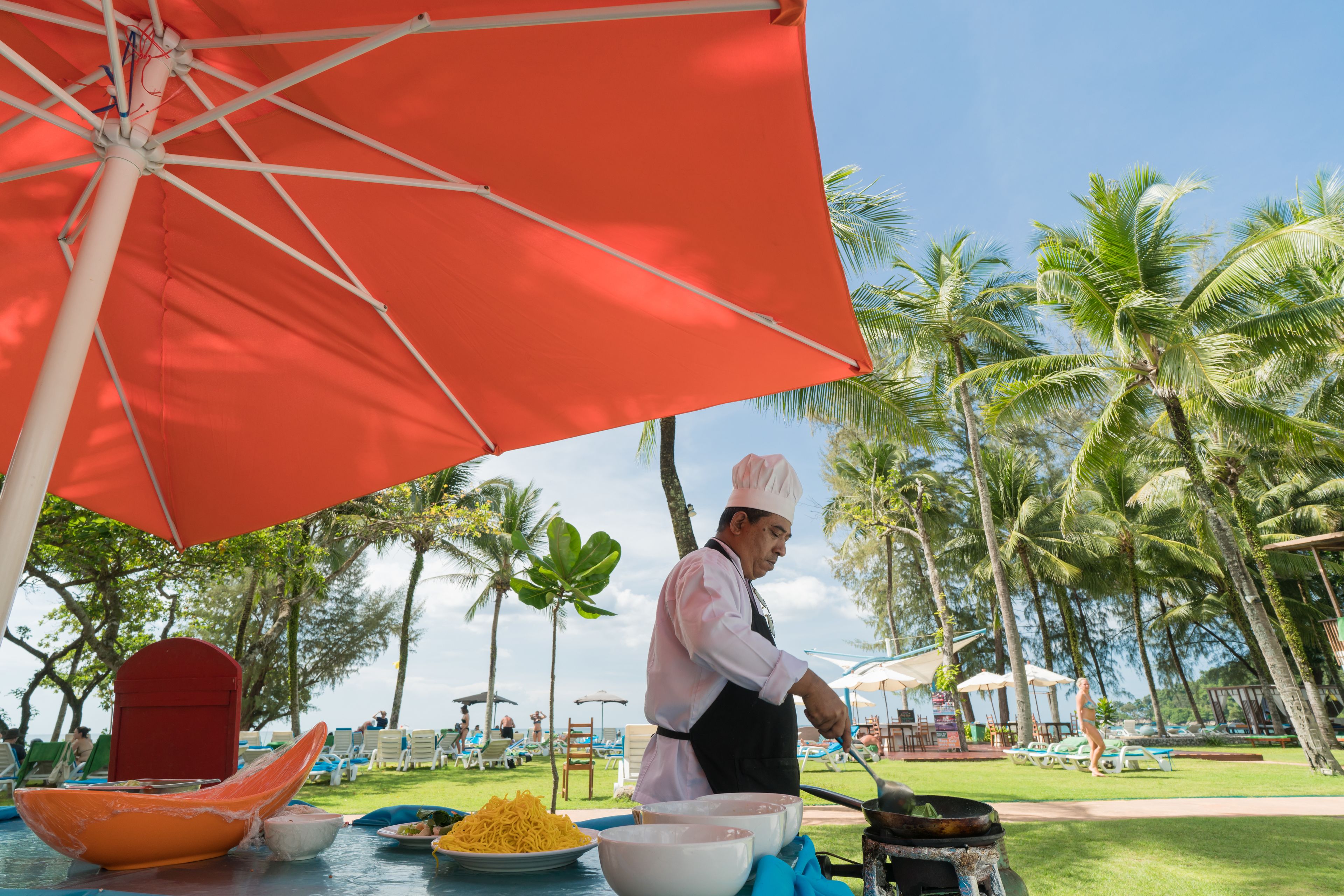 A chef preparing food under a red umbrella at a resort with palm trees and beach chairs in the background.