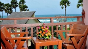 Een houten balkon met twee stoelen en een kleine tafel, met uitzicht op een strand met palmbomen en de oceaan.