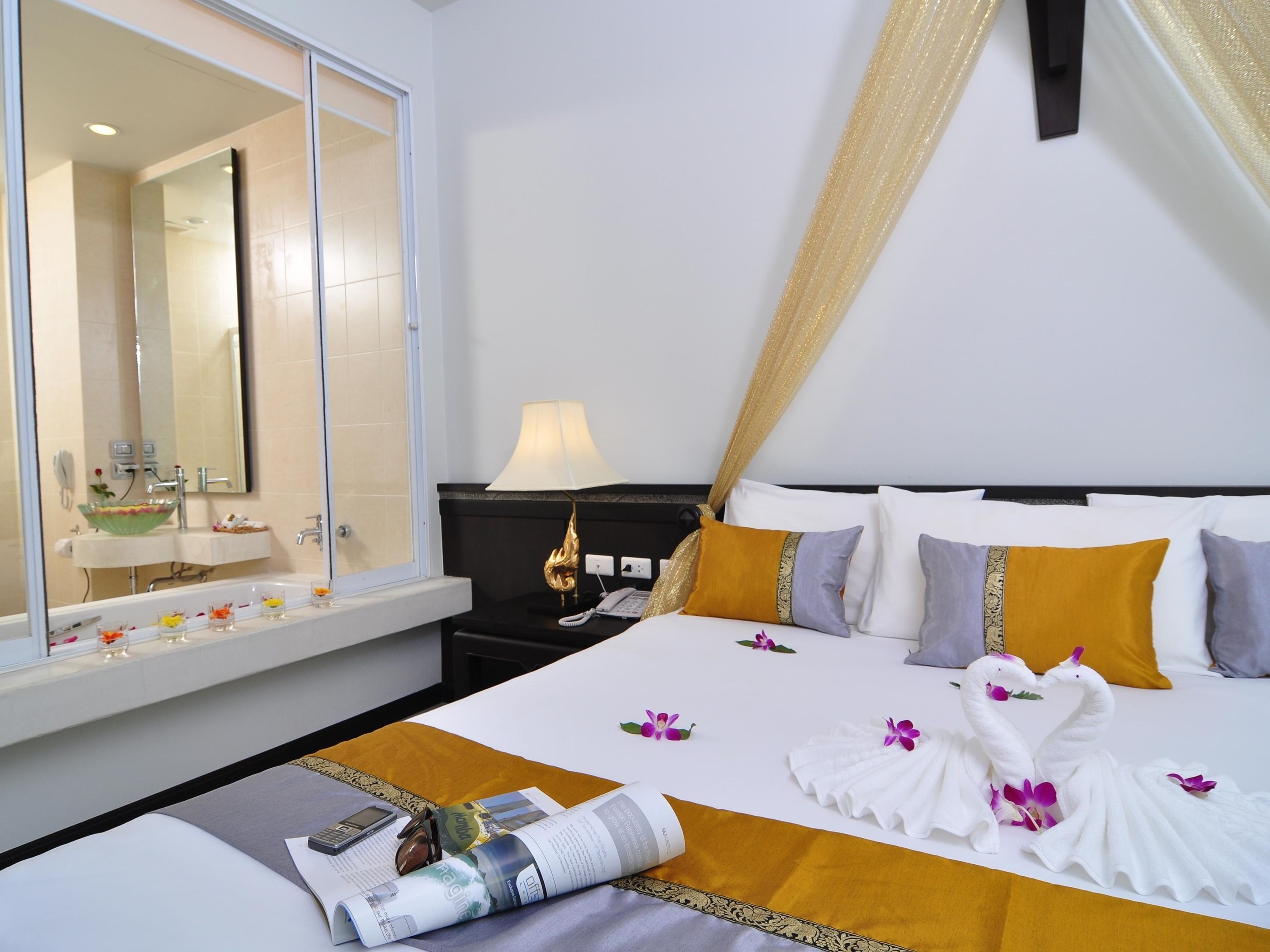 A hotel room with a large bed, a bedside table, a lamp, and a window with a view of the bathroom.
